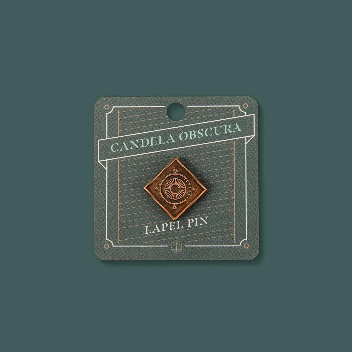 Candela Obscura lapel pin in antique gold with sunburst design and embossed logo.