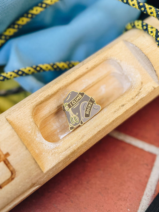 I Climb Rocks Enamel Pin with bold text set in a wooden slot. Rope and blue fabric background visible.