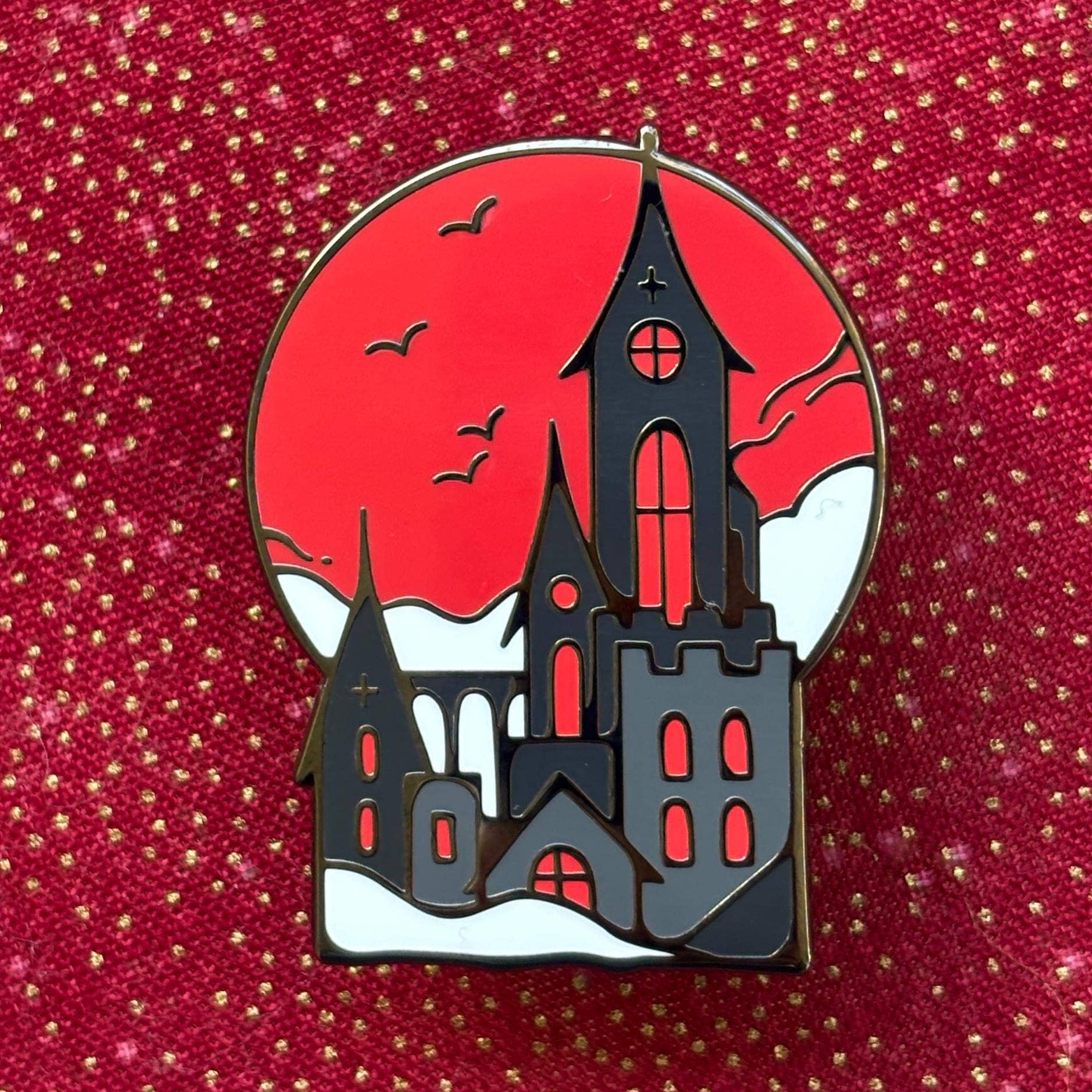 Gothic castle enamel pin with bats, snowy rooftops, red background, and gold plating.