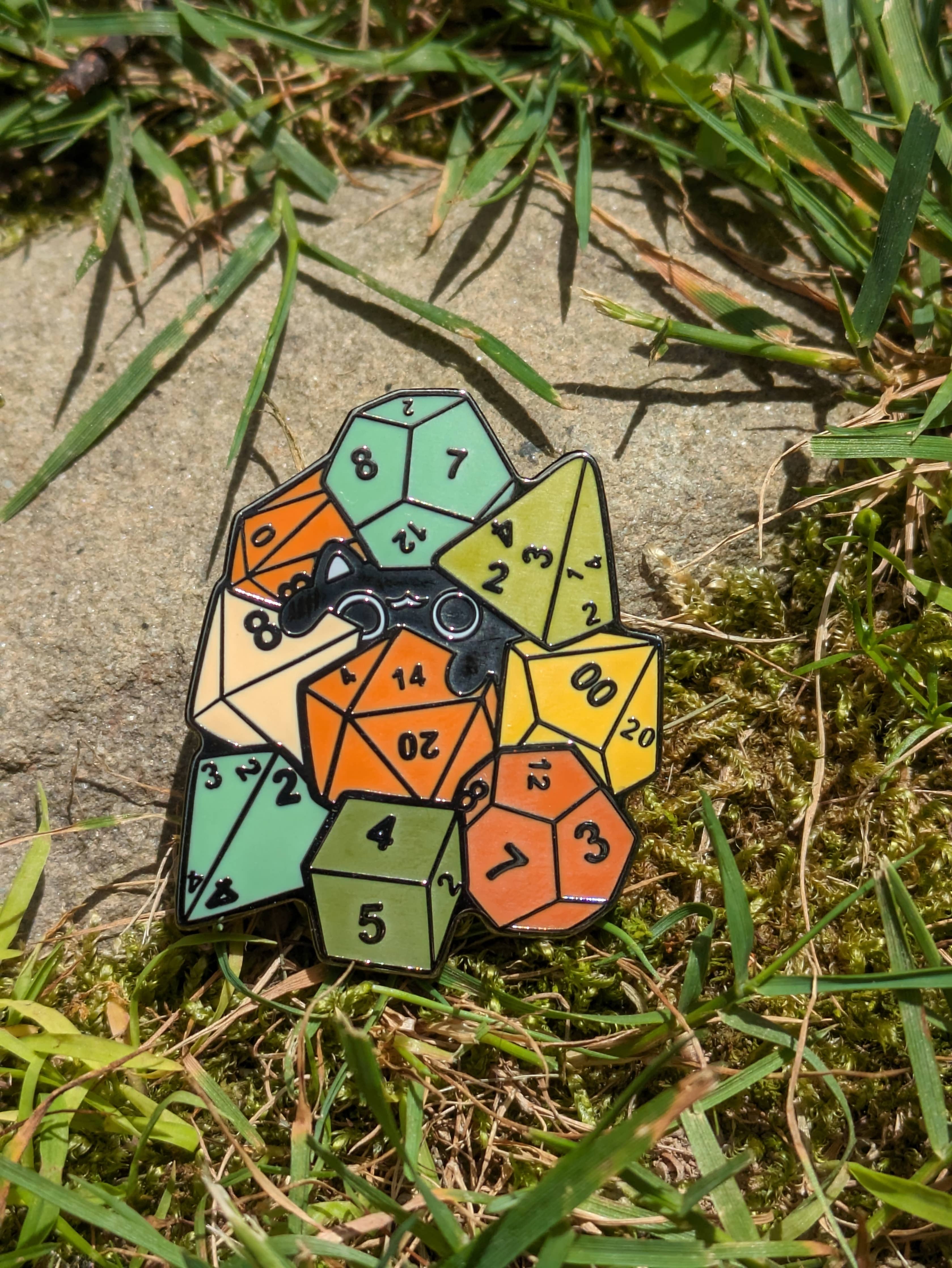 The Cat Amongst Dice Hard Enamel Pin (1.5) with polyhedral dice on grass and stone.