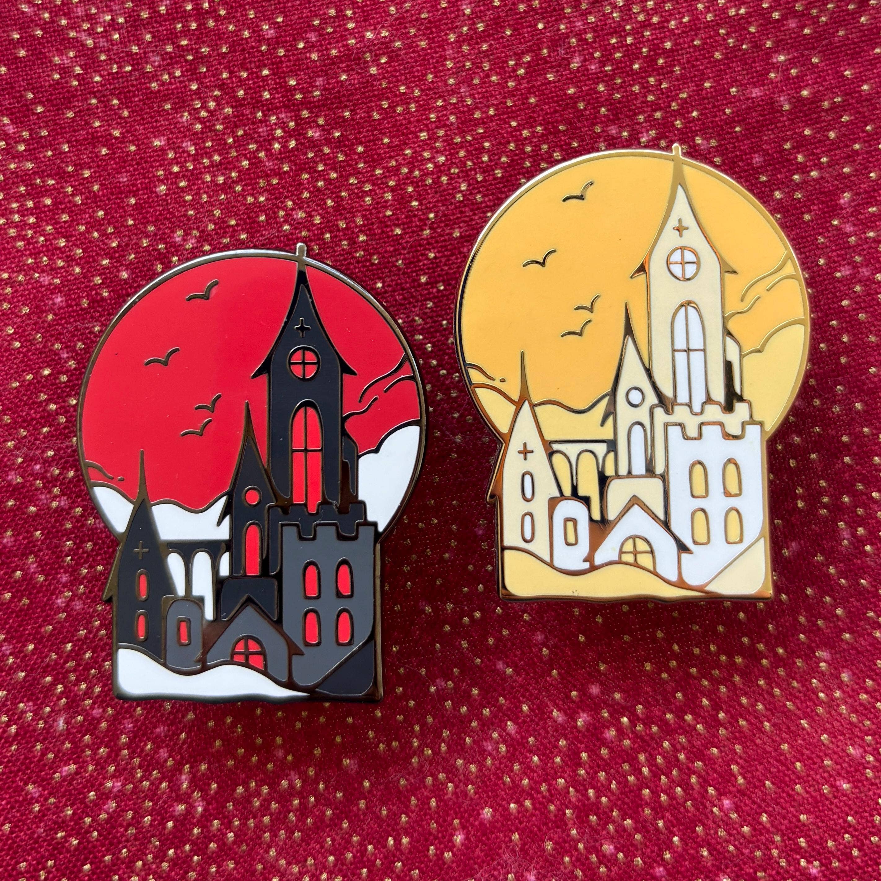 Two castle pins shaped like gothic churches with gold plating; one features a red sky, the other yellow.