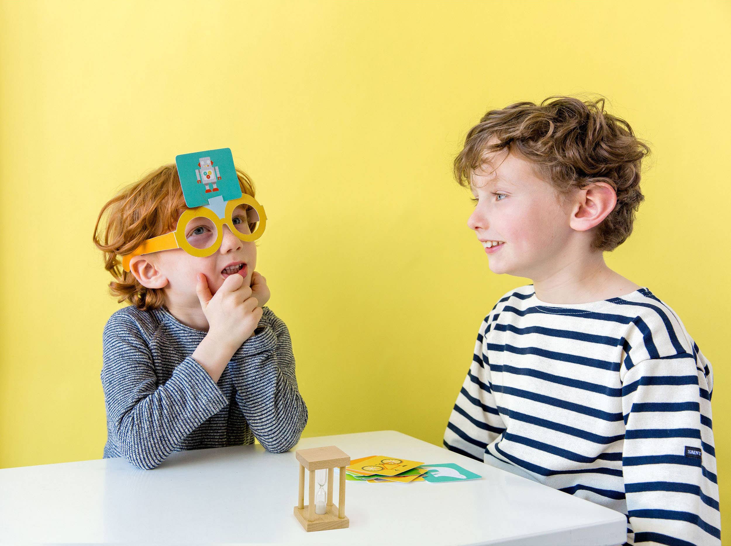 Children play What Am I Game at a table; one child wears glasses with a card on their forehead.