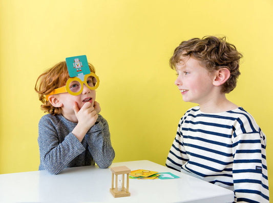 Children play What Am I Game at a table; one child wears glasses with a card on their forehead.