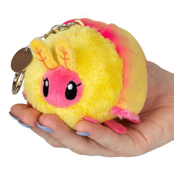 Micro Squishable Rosy Maple Moth keychain, a round plush with large eyes and a metal clip, held in a hand.