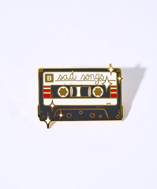 Sad Songs pin: a gold-plated enamel cassette tape with sad songs text, a B label, and a clasp backing.
