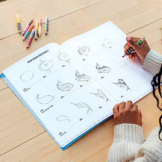 Learn to Draw Under the Sea with Stickers book open on a table with child drawing sea creatures using crayons.