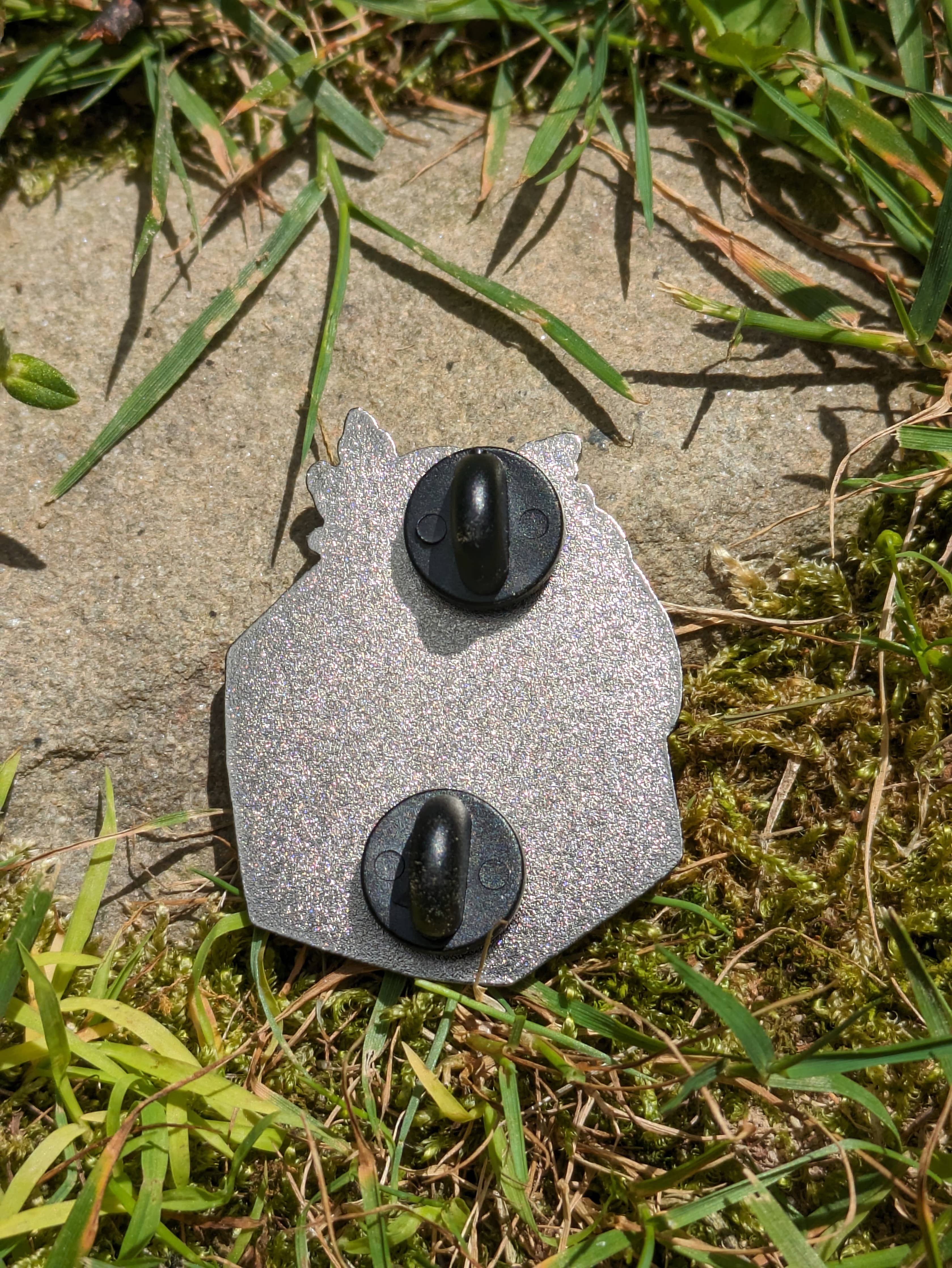 Axolotl on D20 Hard Enamel Pin (1.5) with two black clasps, face down on grass and stone.