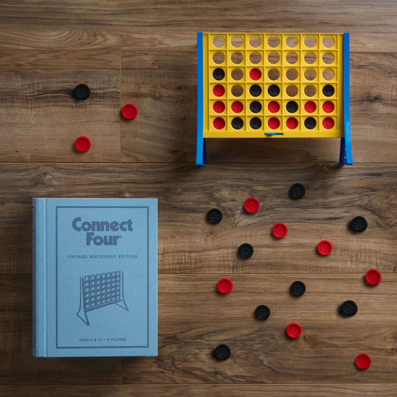Connect Four Vintage Bookshelf Edition with red and black pieces next to the game box on a wood floor.