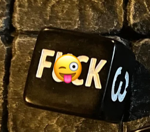 Black F*ck D6 die with white letters, partially covered by a tongue-out emoji on a textured dark surface.