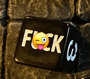 The F*ck D6 is a black gaming die with faces spelling F💋CK, using a tongue-out emoji in place of the U.