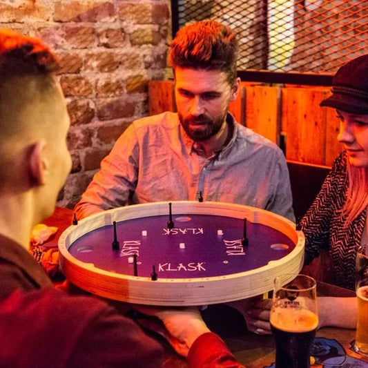 Klask 4 magnetic tabletop game being played by three people at a pub table with drinks nearby.