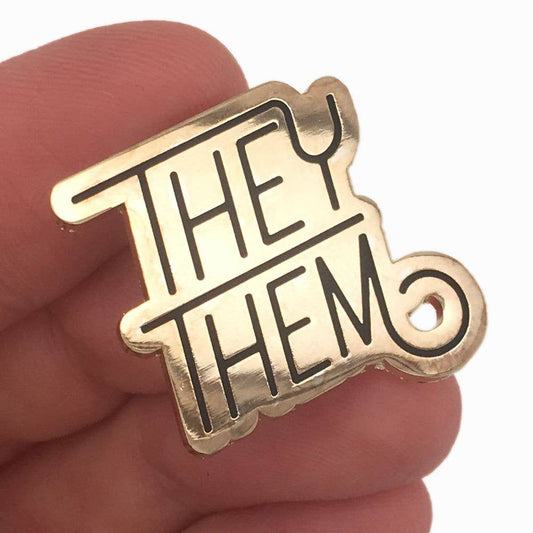 Pronoun Pin - They/Them with bold black letters on a gold background held by a hand.
