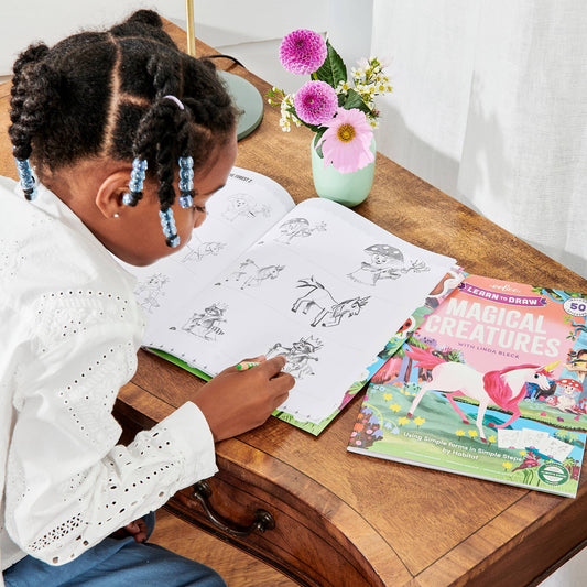 Child drawing in Learn to Draw Magical Creatures with Stickers at a desk; flowers in vase nearby.