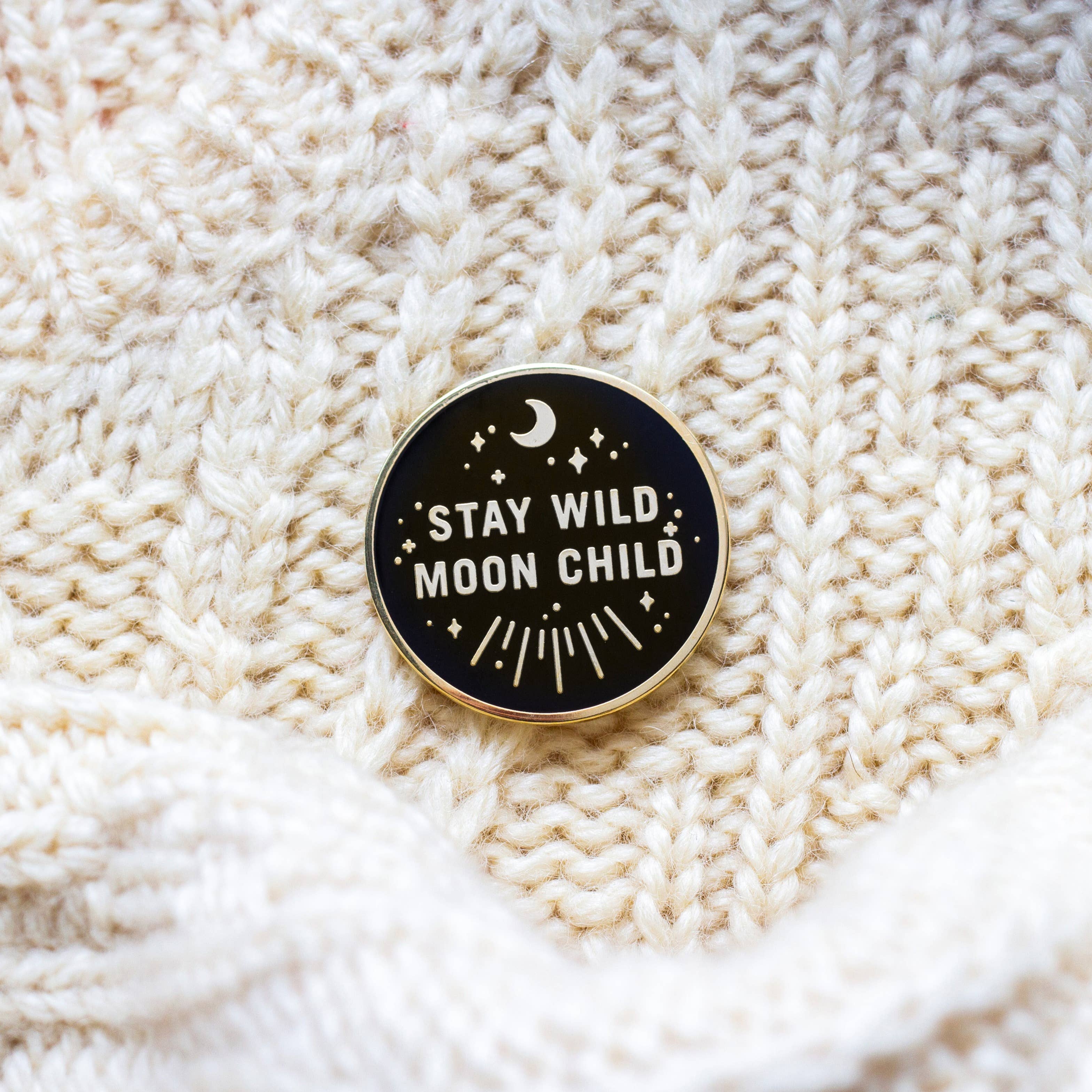 Stay Wild Moon Child enamel pin with celestial designs and the phrase “STAY WILD MOON CHILD” on cream knit background.