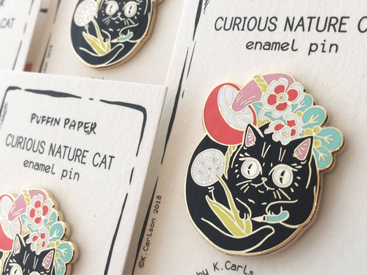 Curious Nature Cat enamel pin: a black cat holding a dandelion surrounded by red, pink, and blue flowers.