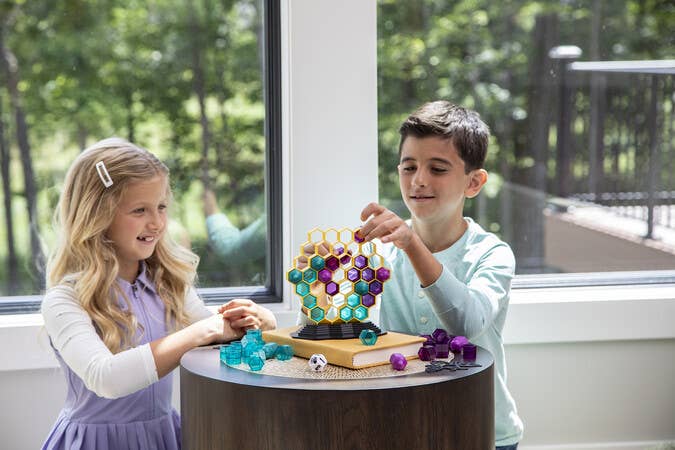Children stack colorful Queen Swarm hexagons by a window. Greenery is visible outside.