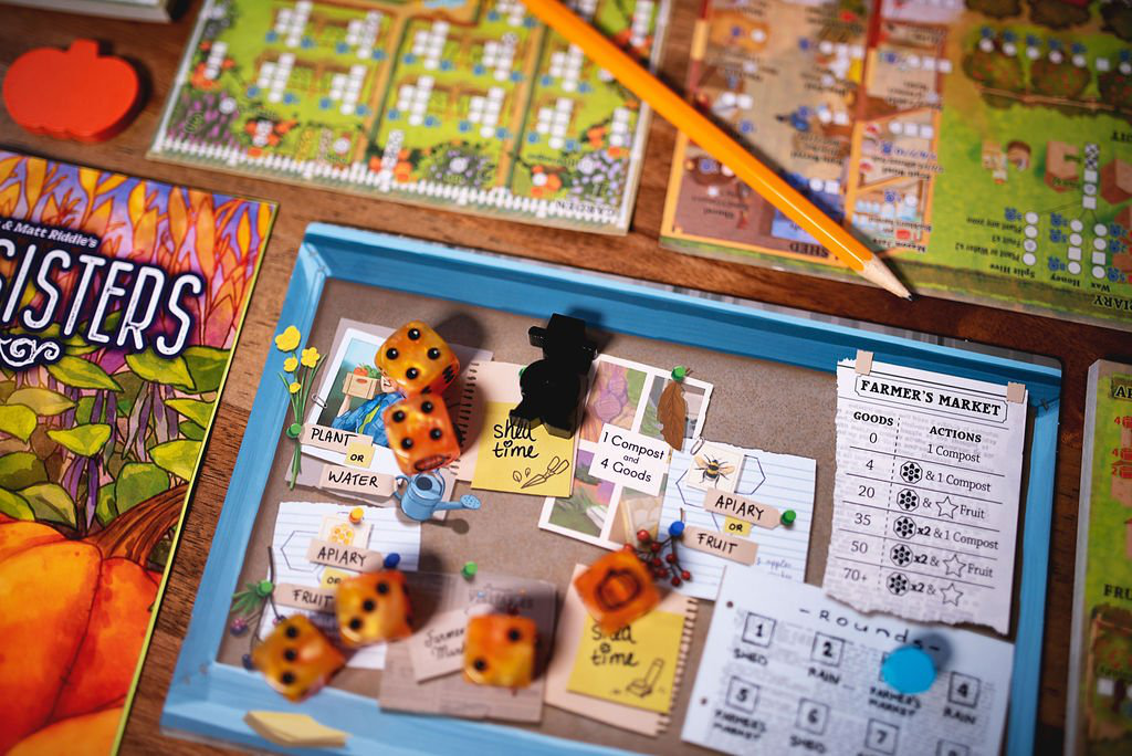 Three Sisters board game components including cards, orange dice, tokens, pencil, and box on a wooden table.