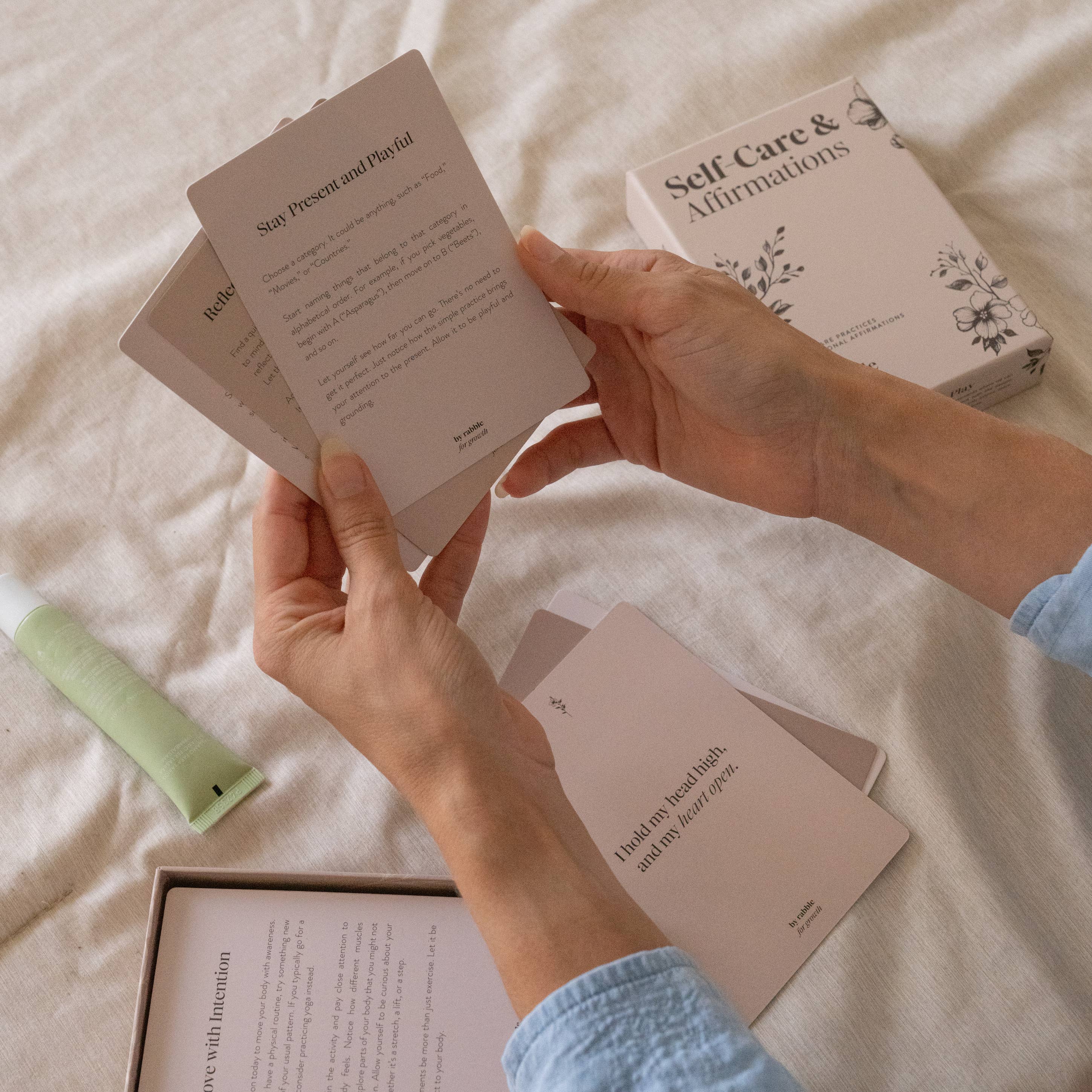 Self-Care & Affirmations cards held in hand with box and green tube on a beige surface.