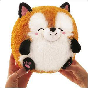 Mini Squishable Baby Fox (7), round plush toy with closed eyes and pink cheeks, held by a person.