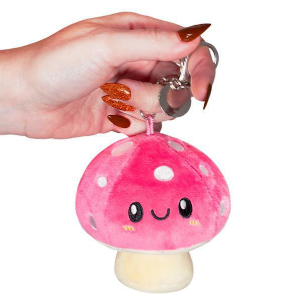 Pink Micro Squishable Mushroom keychain plush with a smiling face and white spots, held in a hand.
