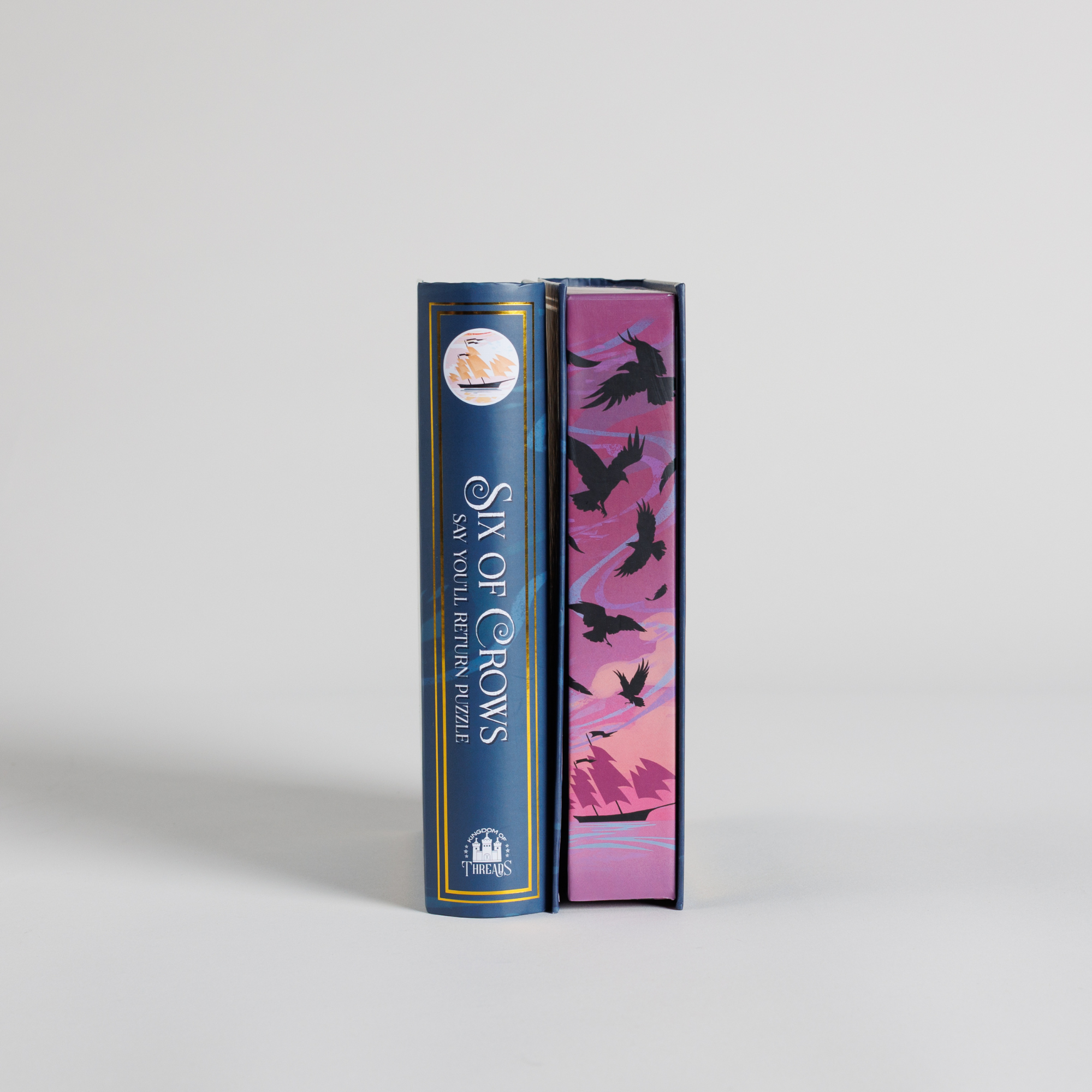 Say You’ll Return Keepsake Puzzle and Six of Crows book spines; black bird design on pink and purple background.