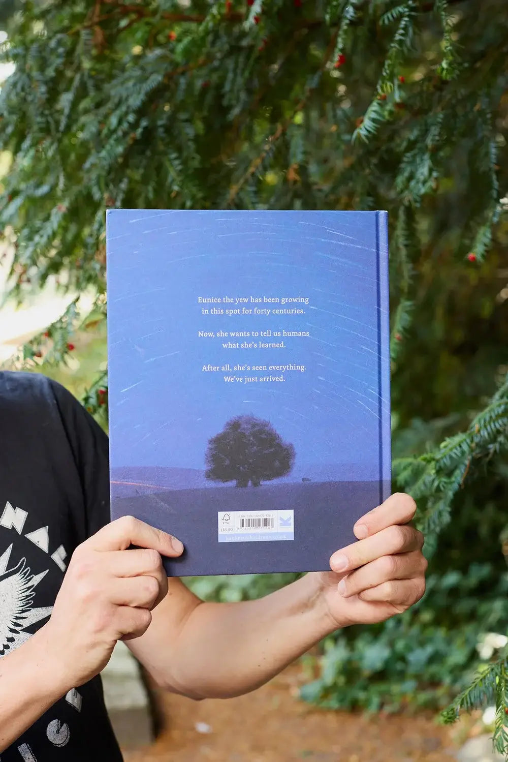 Person holding A Tree is a Time Machine book with tree and starry sky cover; greenery in background.