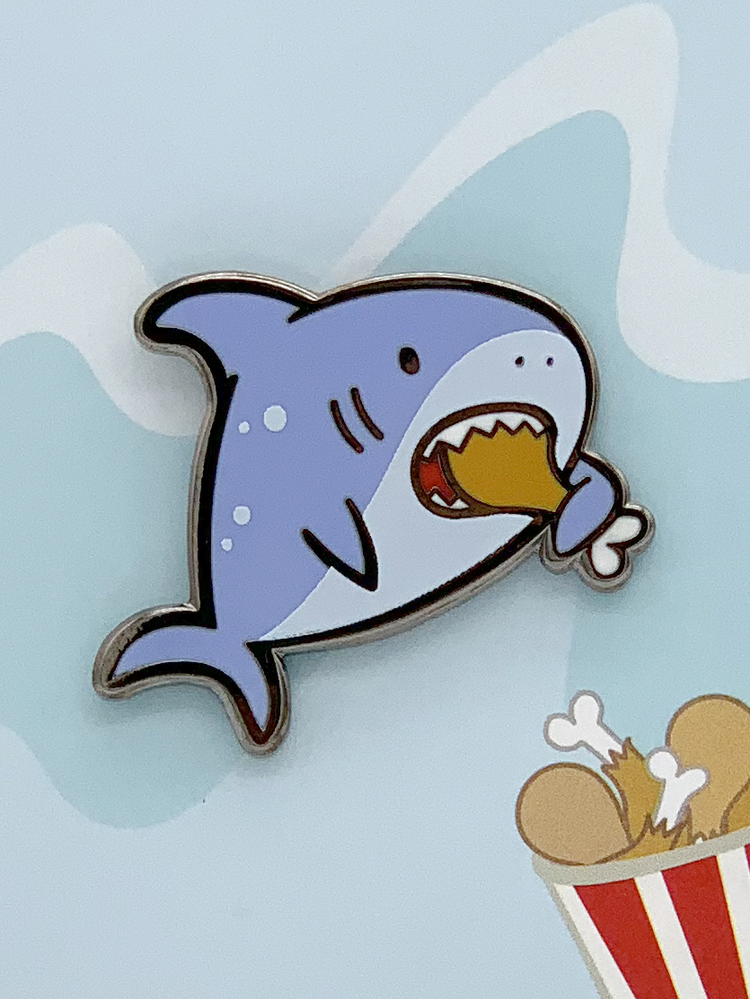 Hungry Shark enamel pin, black nickel plated, shows cartoon shark holding fried chicken and a bucket of chicken legs.