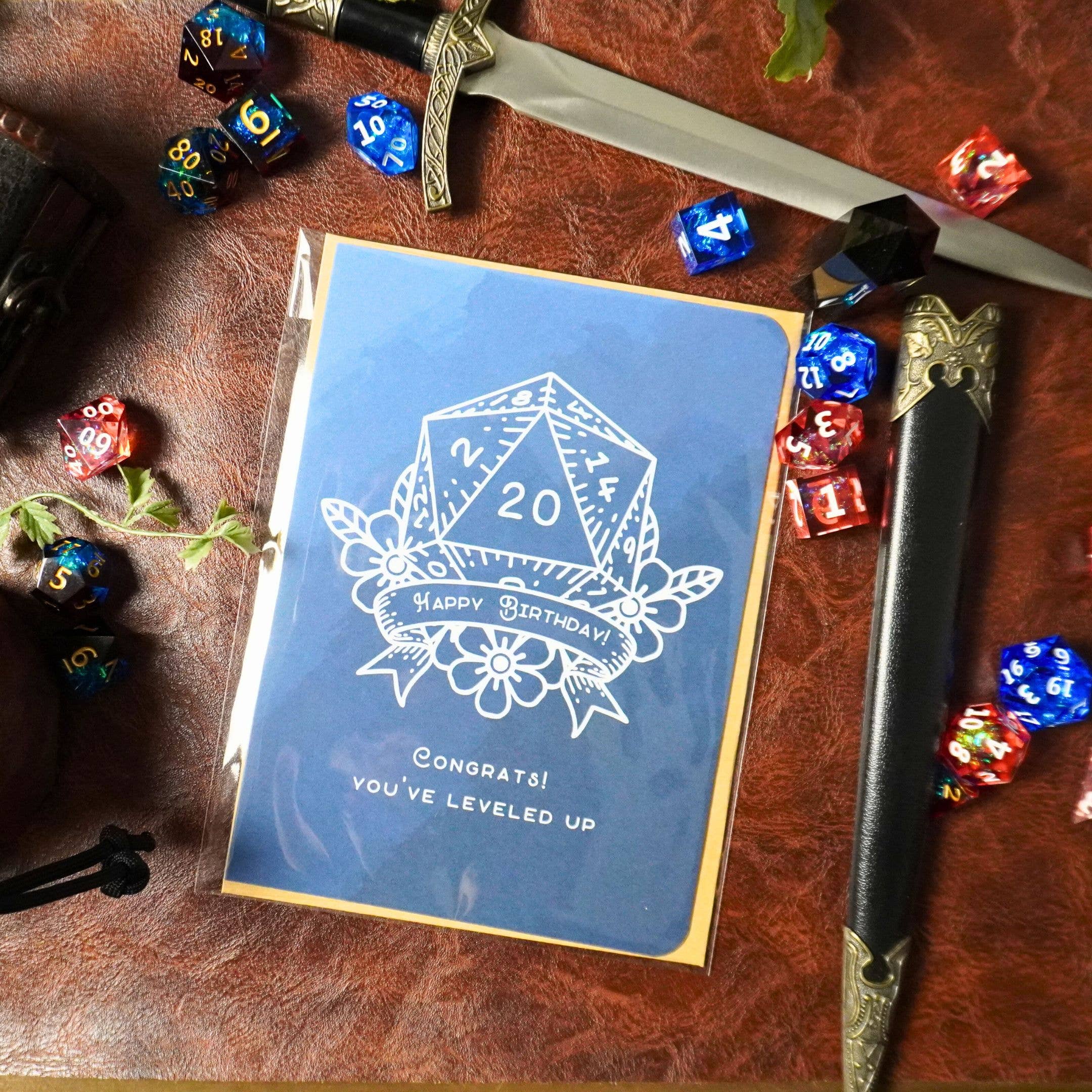 D20 Leveled Up DnD Birthday Card, blue with d20 die, dice, sword, and leather elements.