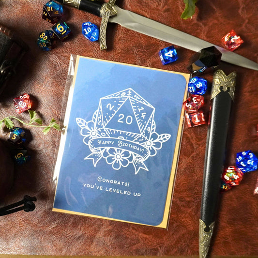 D20 Leveled Up DnD Birthday Card, blue with d20 die, dice, sword, and leather elements.