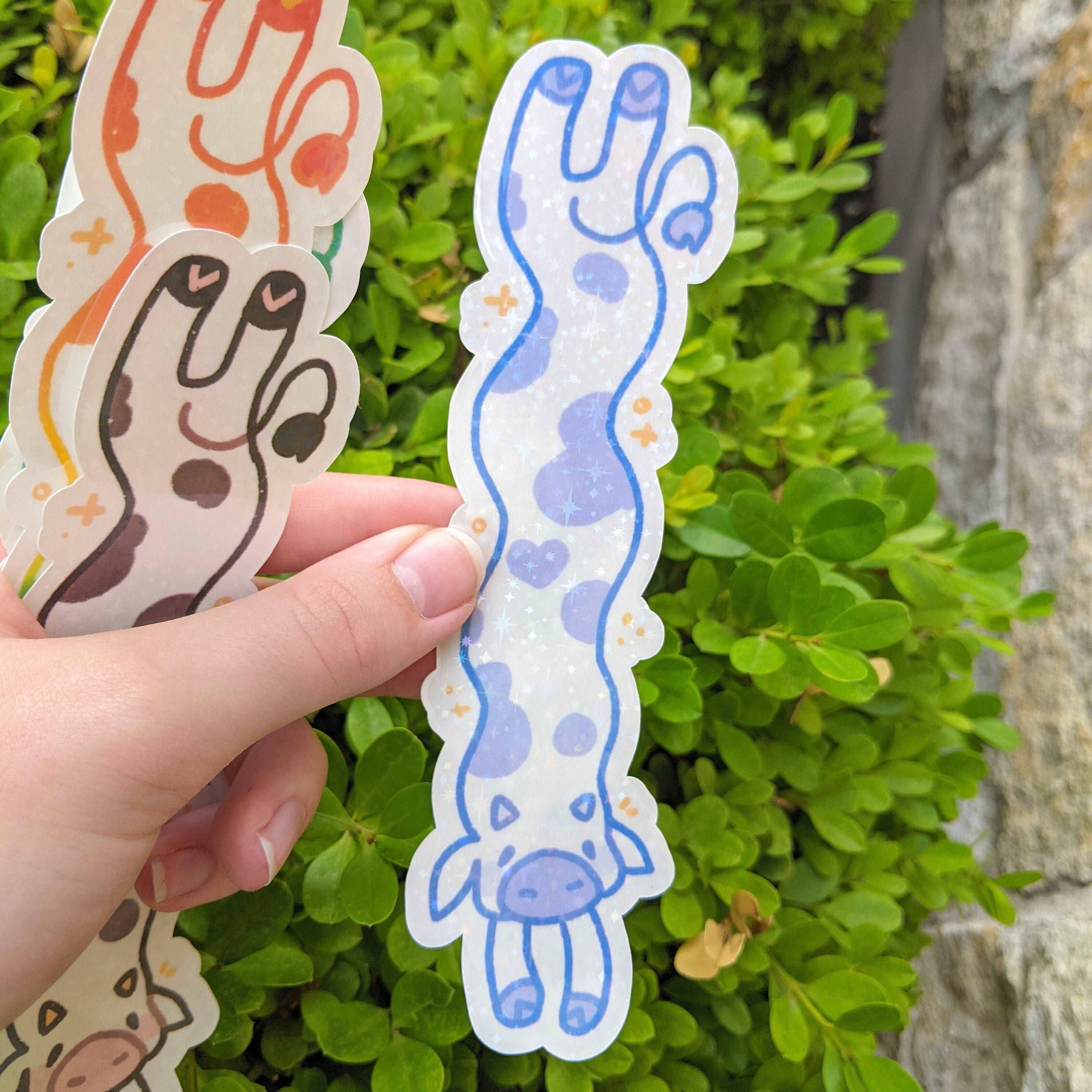 A hand holds a MORE Sparkly Long Cow Sticker next to a green bush.