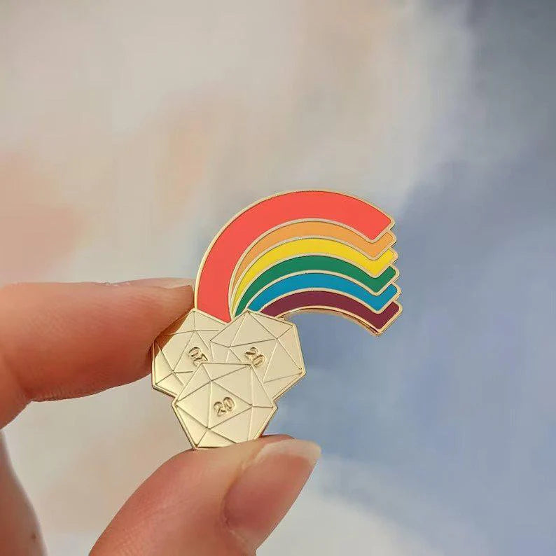 End of the Rainbow Hard Enamel Pin featuring a rainbow above three gold 20-sided dice, held in a hand.