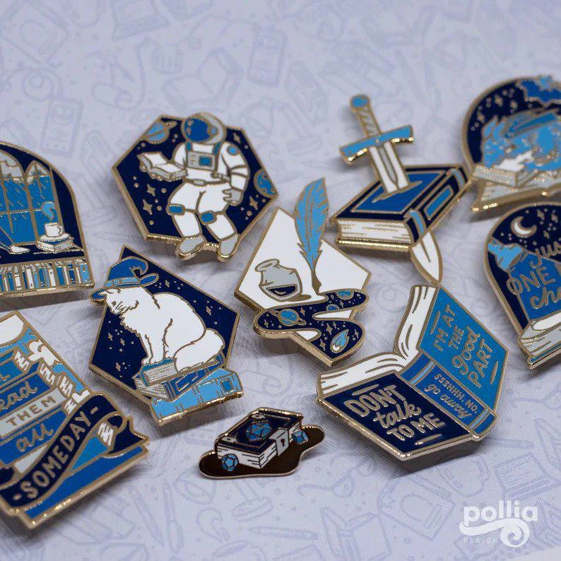 Gold-plated Booxcalibur enamel pin with books, an astronaut, a polar bear, and fantasy elements.