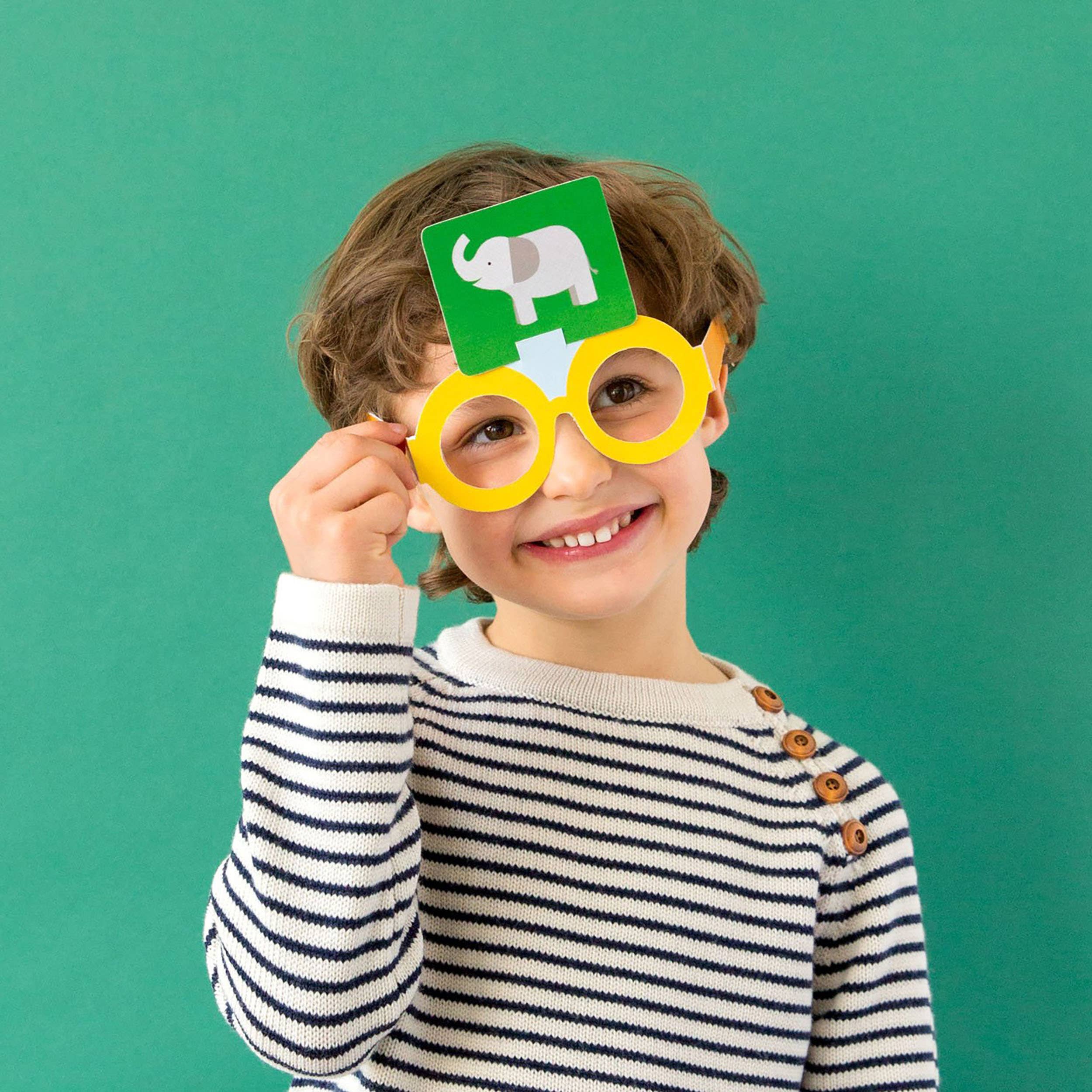 Child playing What Am I Game, holding elephant card to forehead, wearing yellow glasses.