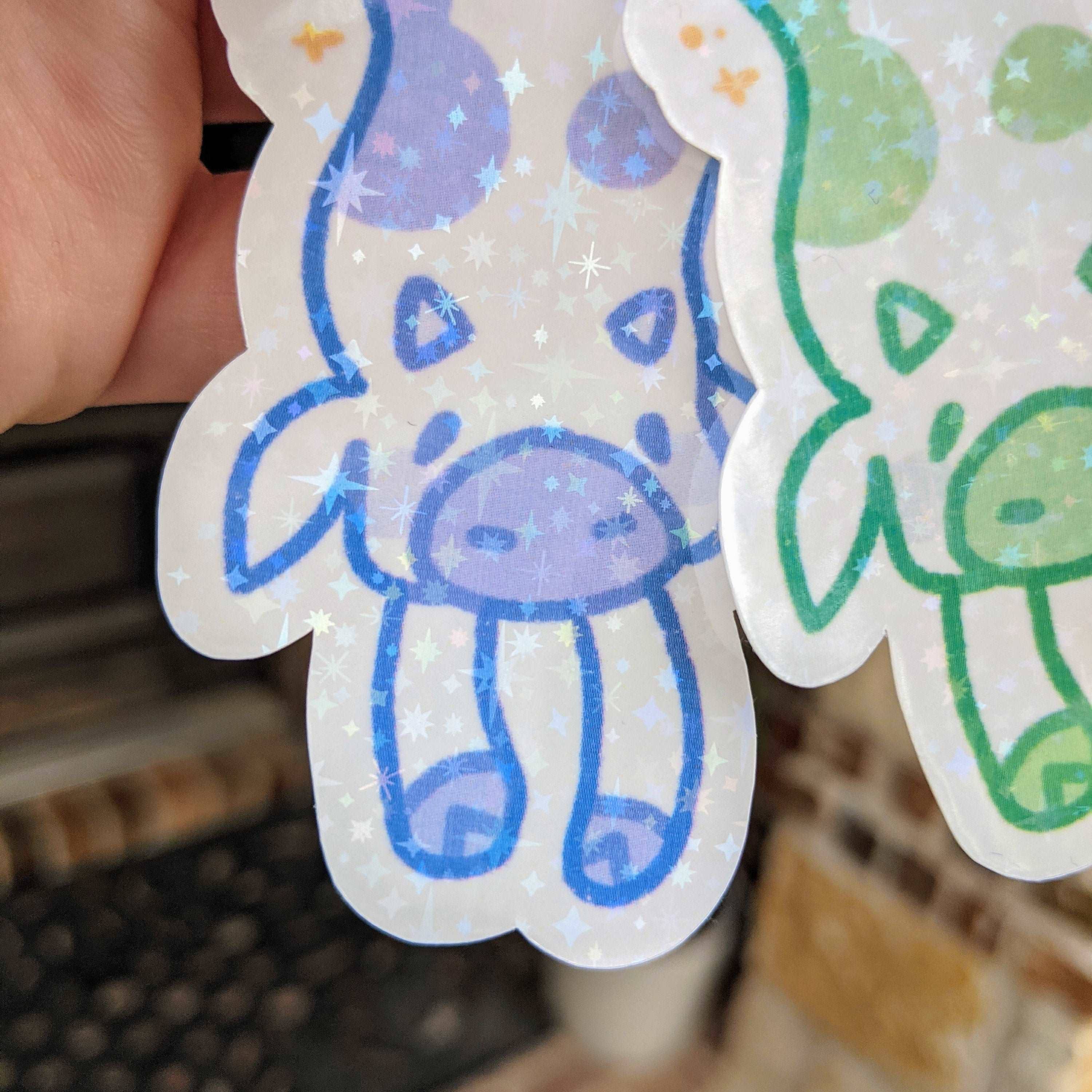 Hand holding two MORE Sparkly Long Cow Stickers in blue-purple and green; both are waterproof.