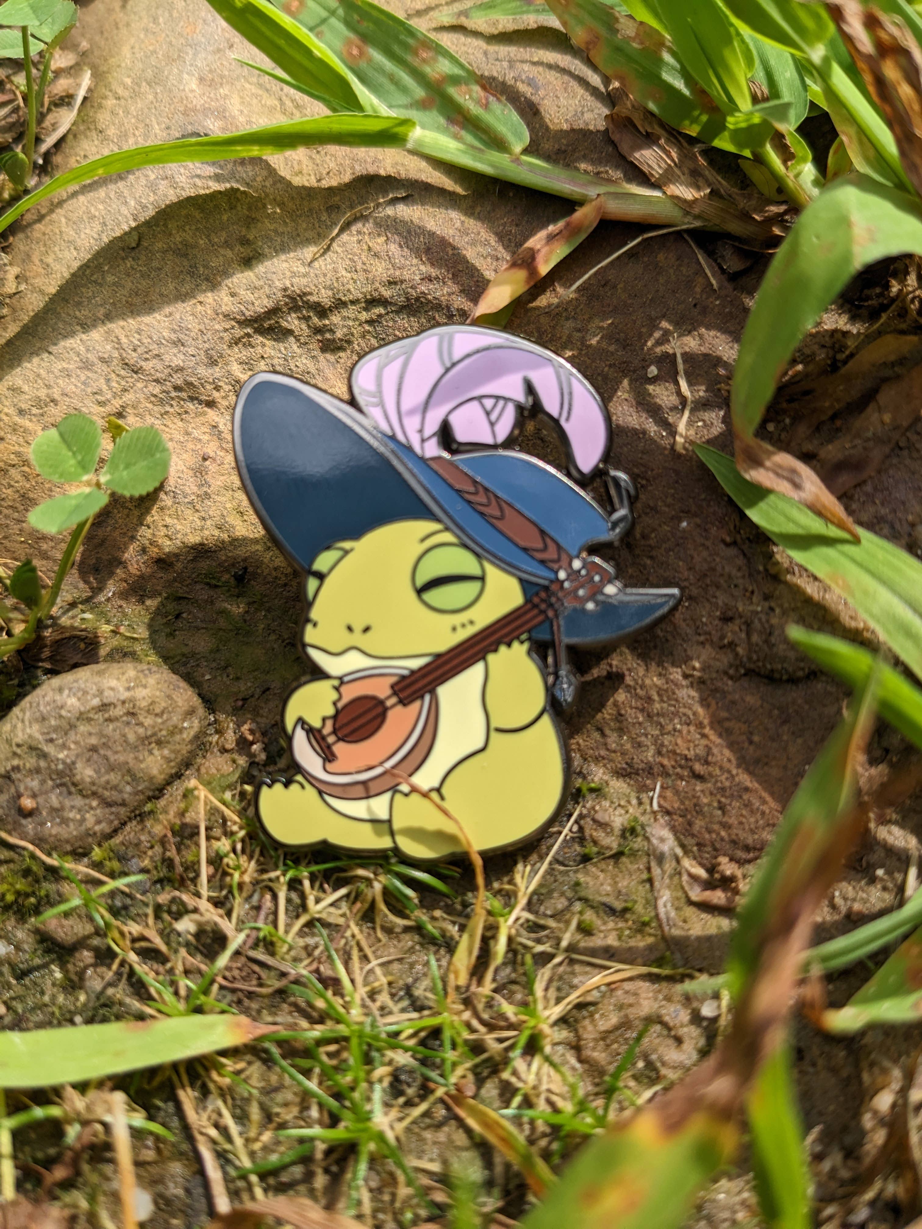 1.5 enamel pin featuring a frog bard in a blue hat playing a lute.