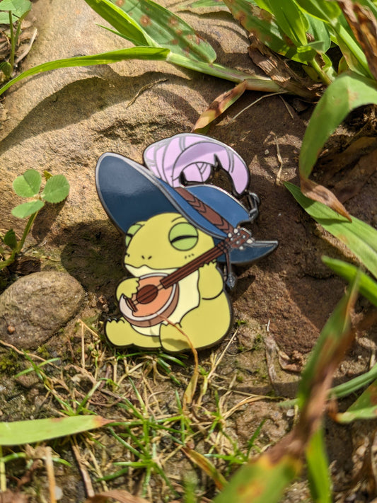 1.5 enamel pin featuring a frog bard in a blue hat playing a lute.