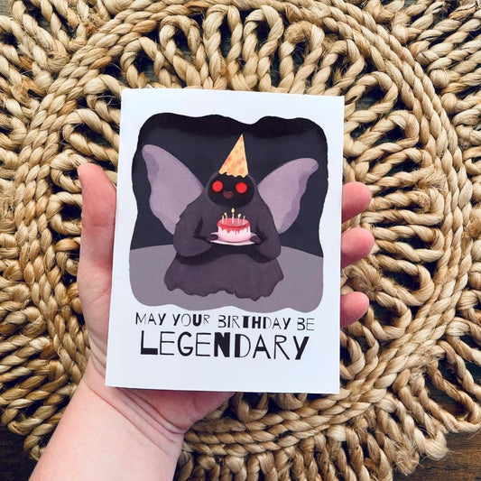 Mothman Cryptid Funny Birthday Card held in hand, features Mothman with cake and party hat. Text: May your birthday be legendary.