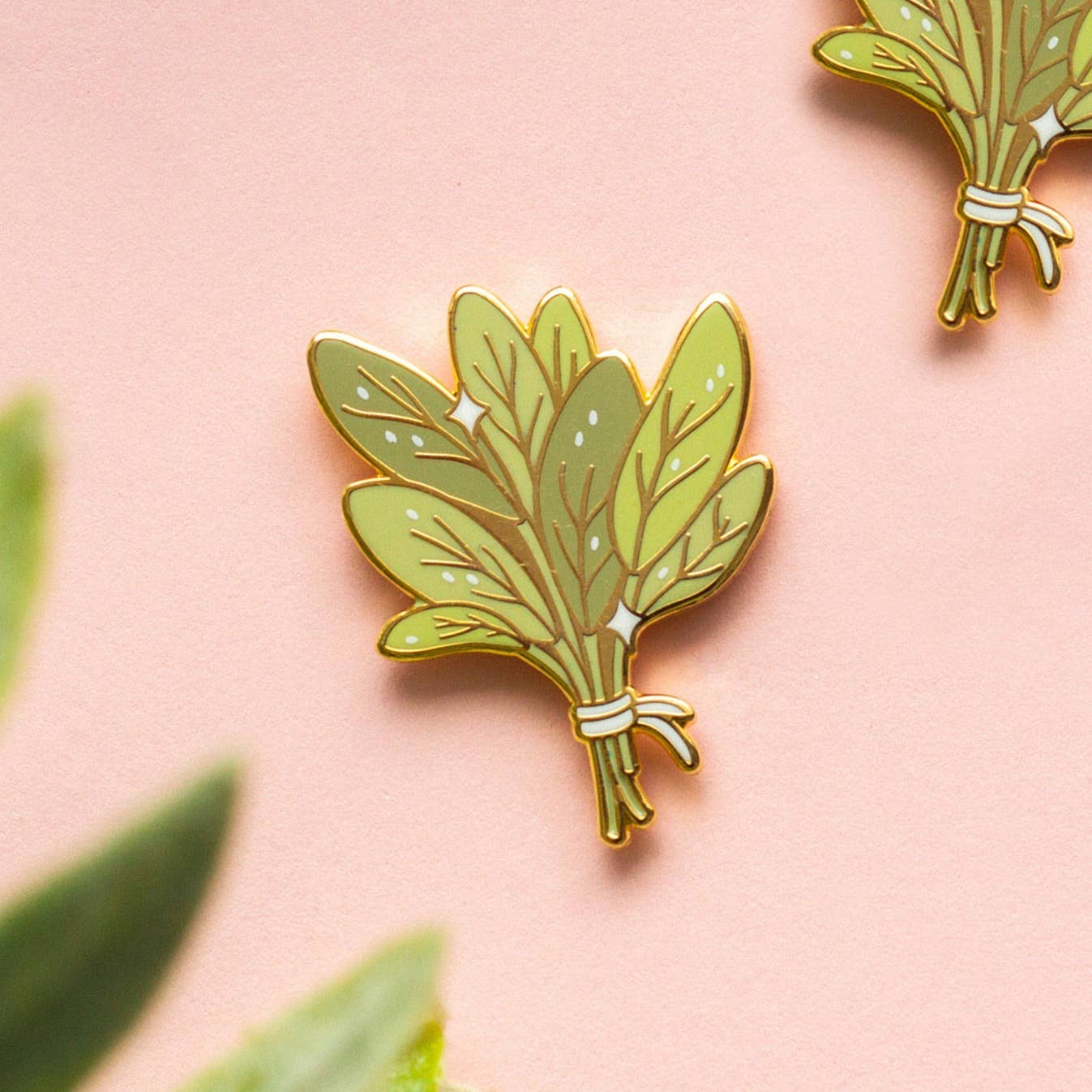 Bundle of Sage Enamel Pin with gold-outlined green herbs on a pale pink background.