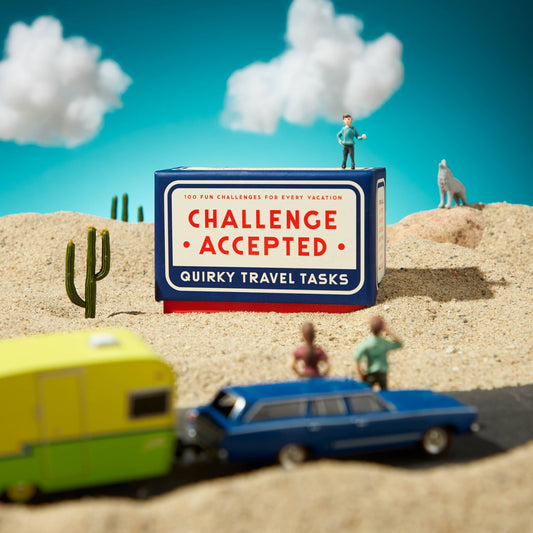 Brass Monkey Challenge Accepted Travel Tasks Card Deck on sand with toy cars, cards, figures, cactus, and wolf.