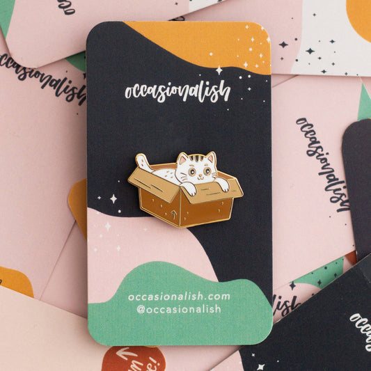 Box Cat Enamel Pin in a box, displayed on a card with occasionalish branding.