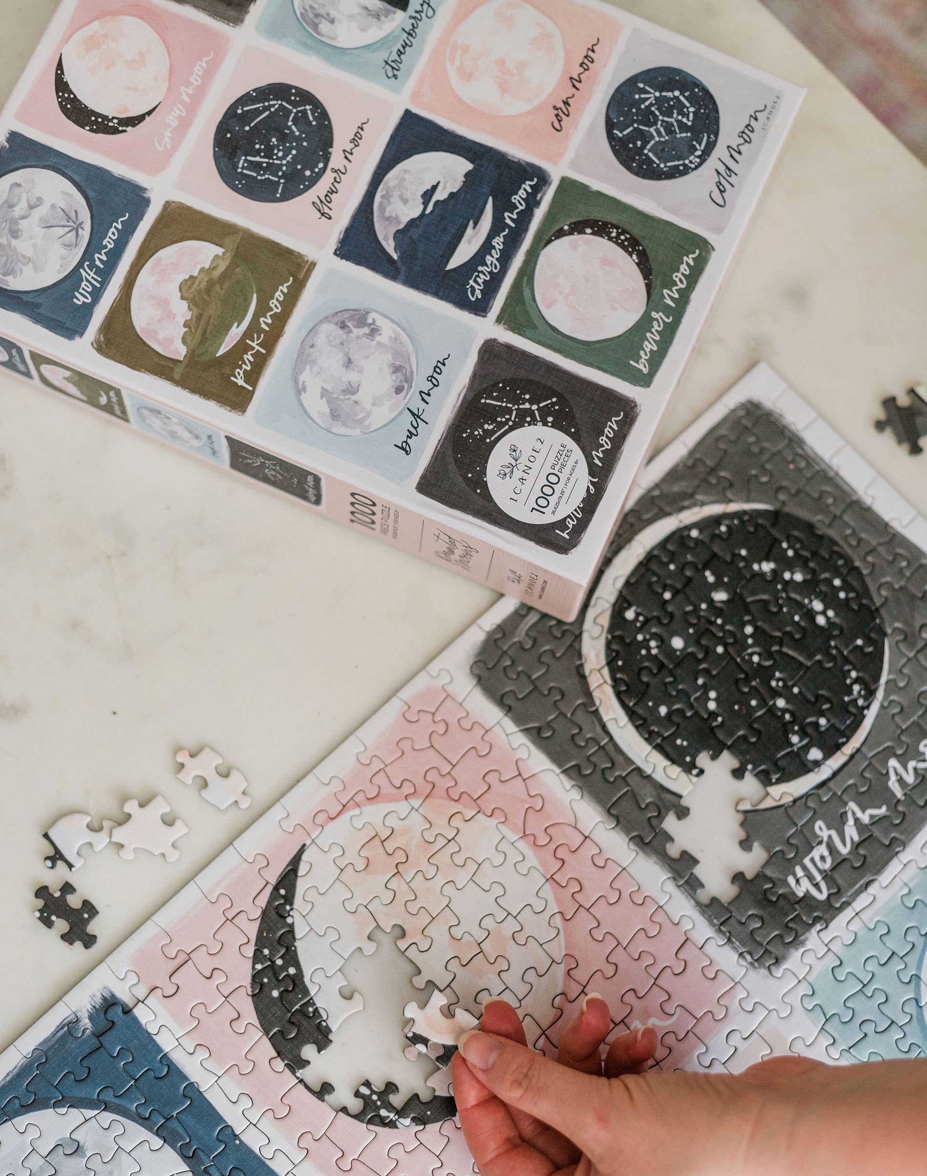 Hand places a piece in the Painted Moons 1,000 Piece Jigsaw Puzzle beside its box on a white surface.