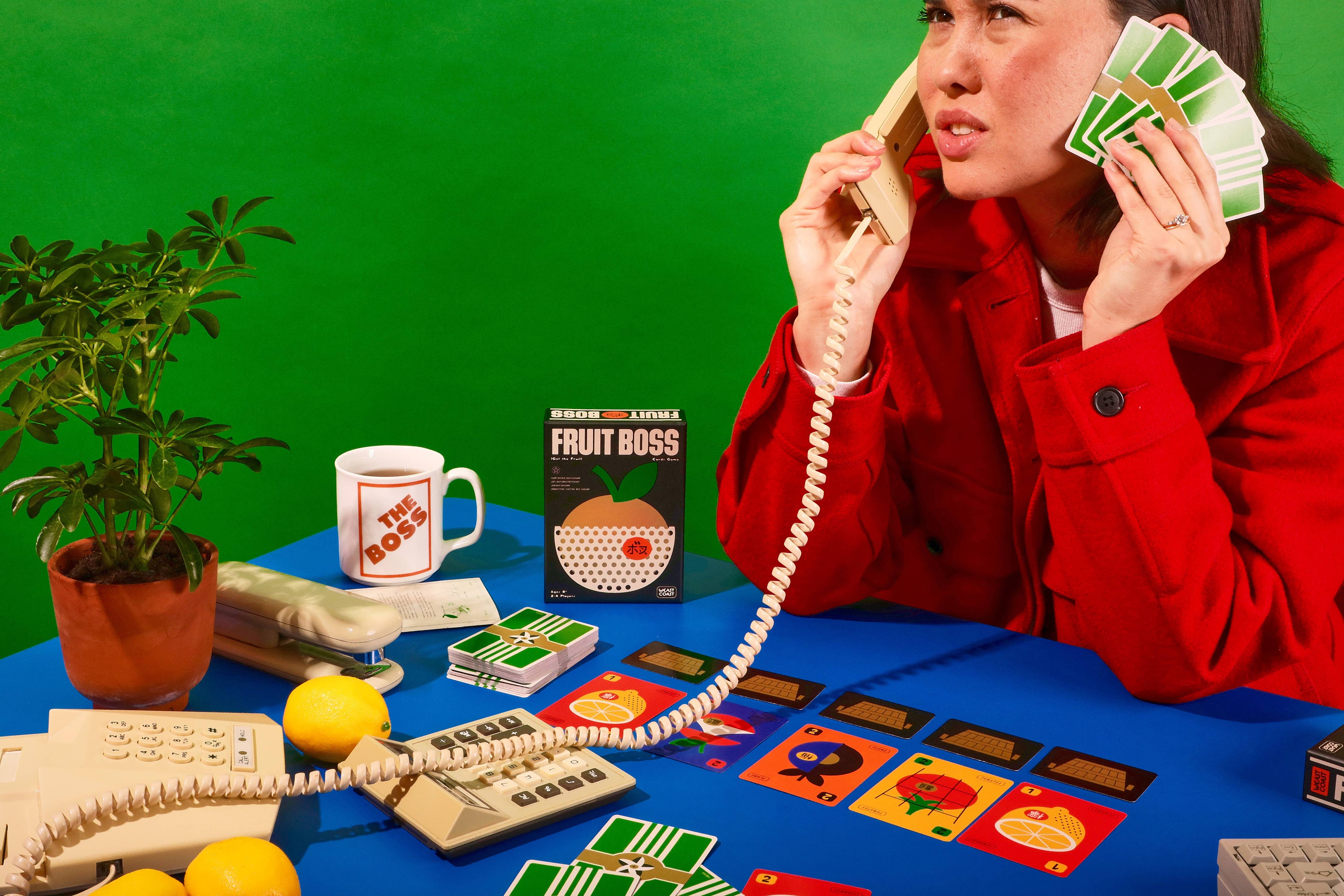 Woman in red coat using Fruit Boss at a cluttered desk with phones, lemons, and potted plant.