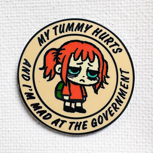 Round black metal enamel pin featuring a messy-haired character and the text, My tummy hurts and I’m mad.