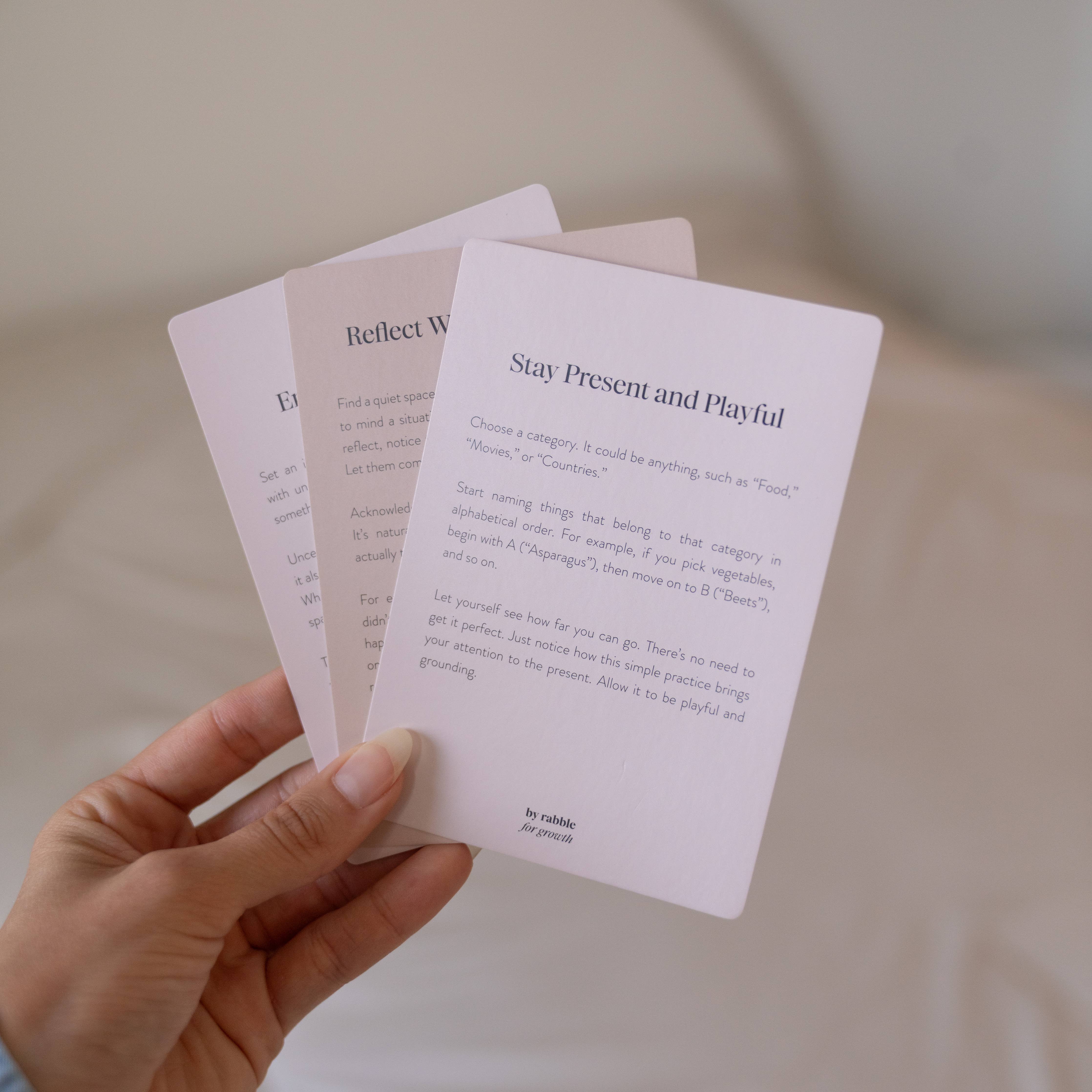 Three Self-Care & Affirmations cards held in a hand; top card reads Stay Present and Playful.