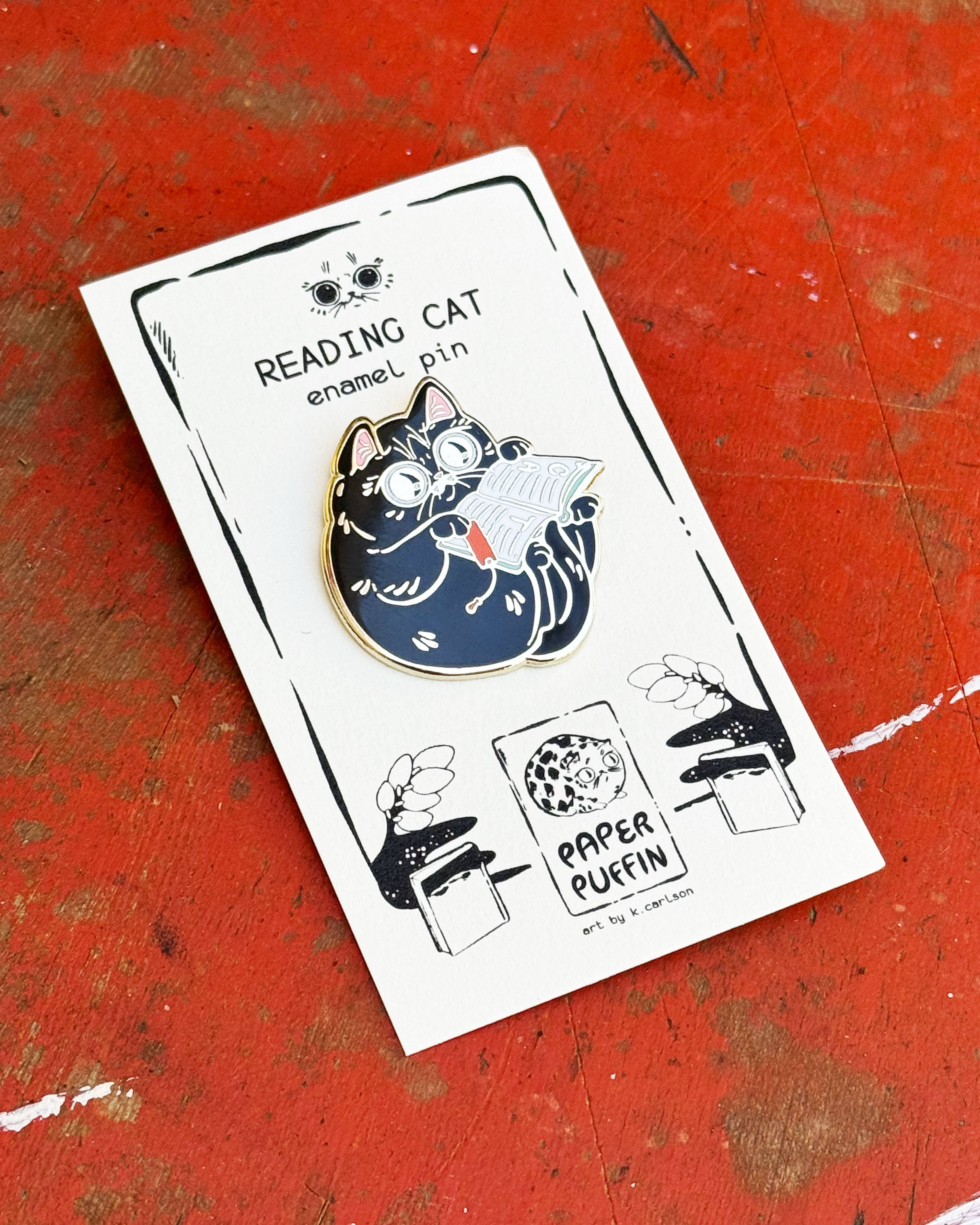 Book Reader Cat enamel pin: a black cat wearing glasses reading an open book.
