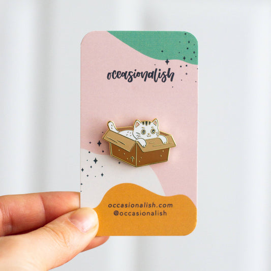 Box Cat Enamel Pin by Occasionalish held in a hand inside a gift box.