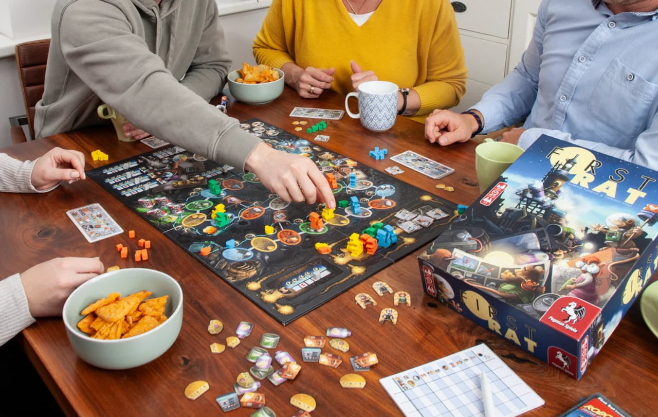 Four people playing First Rat board game at a table with game pieces, snacks, and drinks.