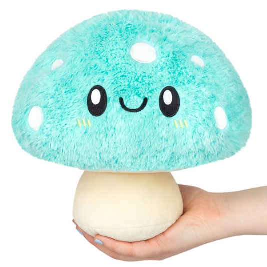 A hand holds the Turquoise Mushroom Squishable plush, featuring a smiling face.