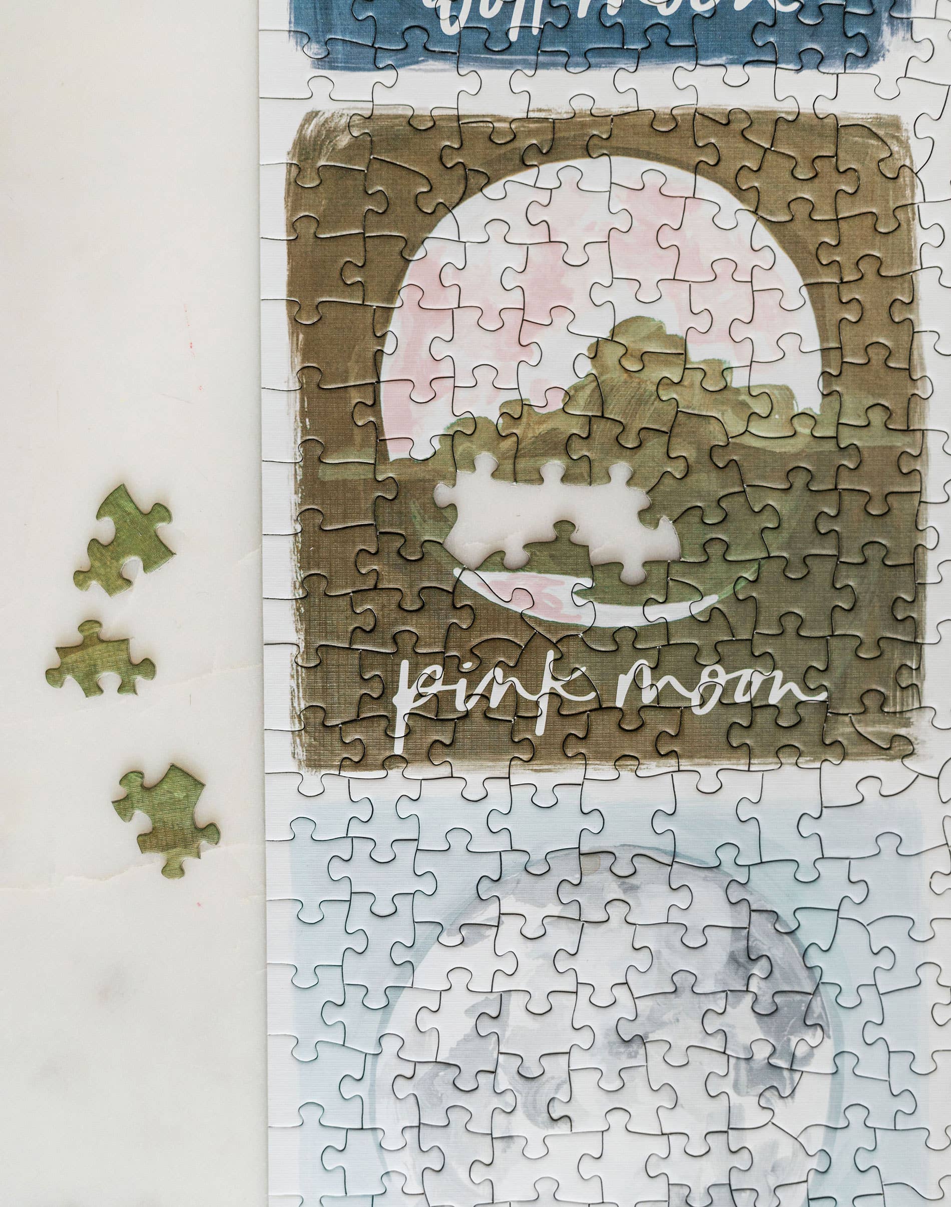 The Painted Moons 1,000 Piece Jigsaw Puzzle with painted landscape, three missing pieces, and the words Pink Moon.