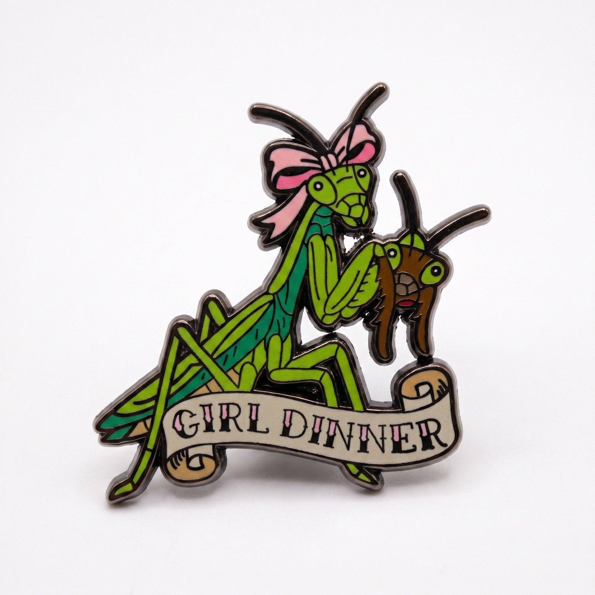 Girl Dinner Mantis pin: green mantis in a pink bow holding a bug above a banner, with black plating.